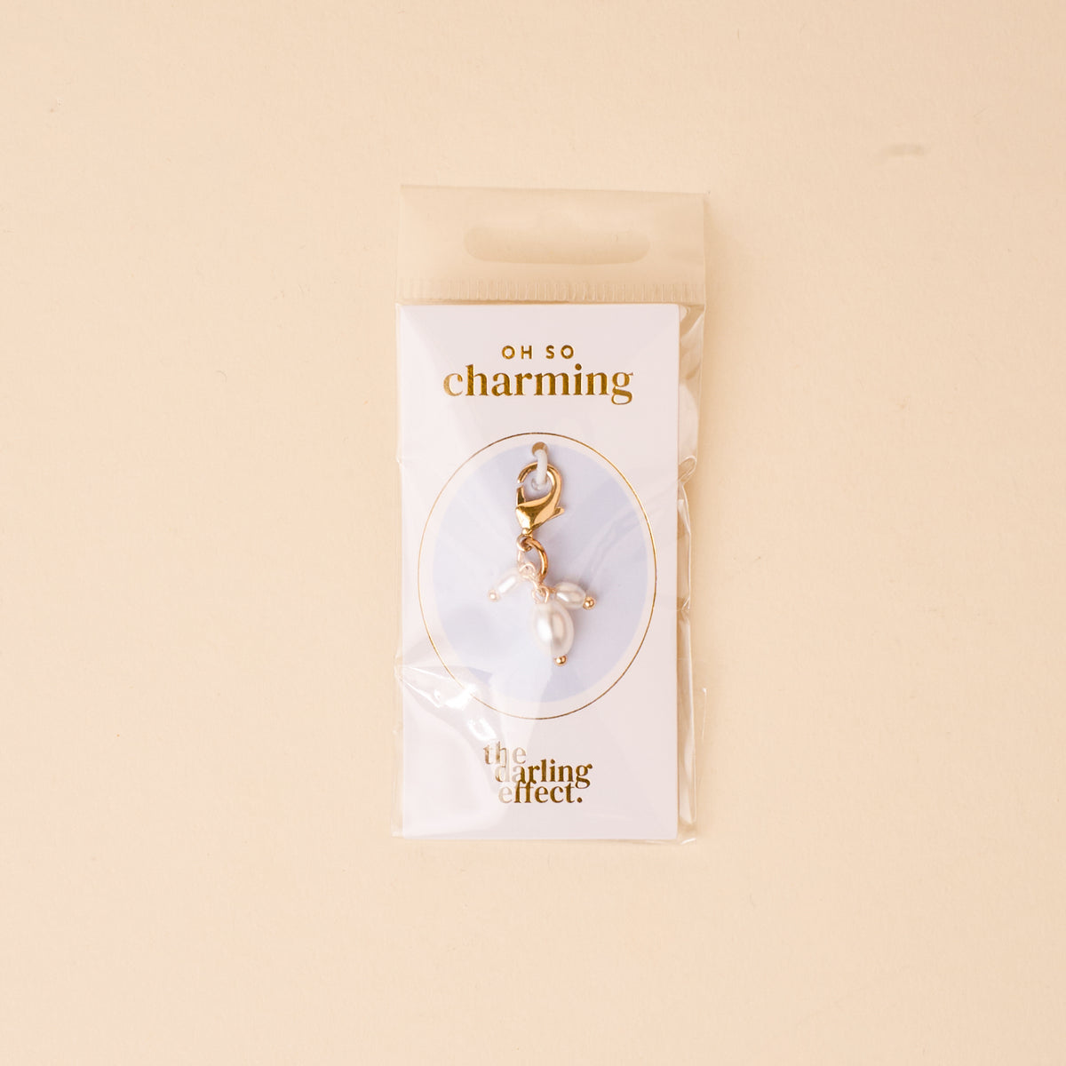 The Pearl Trio Jewelry Charm Gold by the-darling-effect comes in a clear pouch on a card with "OH SO charming" and "the darling effect." The soft beige background makes it perfect for any charm bar collection.
