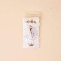 The Pearl Trio Jewelry Charm Gold by the-darling-effect comes in a clear pouch on a card with "OH SO charming" and "the darling effect." The soft beige background makes it perfect for any charm bar collection.