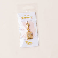 The R Jewelry Charm Initial Letter Gold by the-darling-effect features a gold-plated rectangular pendant with an 'R' in crystals, surrounded by sparkling stones and a lobster clasp. Packaged with signature Oh So Charming branding.