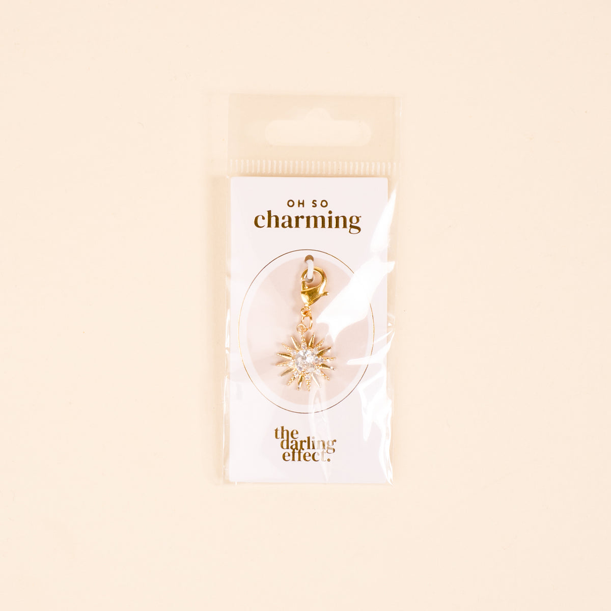 The Star Burst Jewelry Charm Gold by the-darling-effect features a gold star with clear gems on a lobster clasp, perfect for your charm bar. Packaged on a white card with OH SO charming and the-darling-effect logos—timeless elegance for your collection.