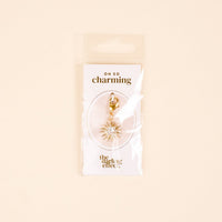 The Star Burst Jewelry Charm Gold by the-darling-effect features a gold star with clear gems on a lobster clasp, perfect for your charm bar. Packaged on a white card with OH SO charming and the-darling-effect logos—timeless elegance for your collection.