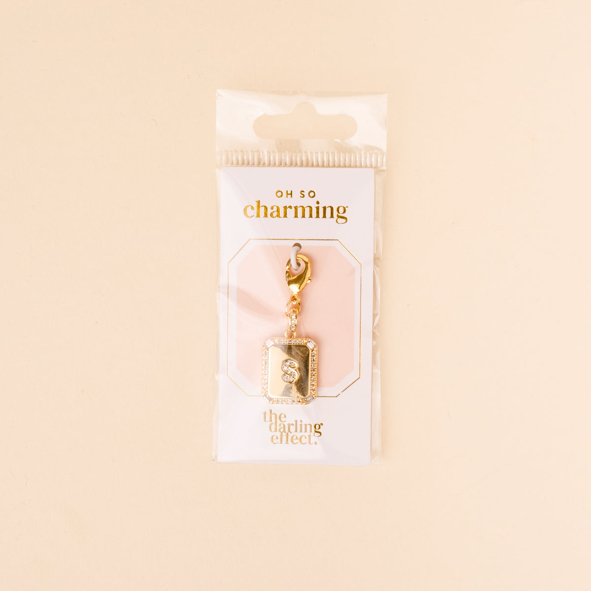 The-darling-effect S Jewelry Charm Initial Letter Gold, a gold-plated brass keychain with a rectangular pendant, is displayed in clear Oh So Charming packaging—an elegant, gift-ready piece set against a soft beige background.