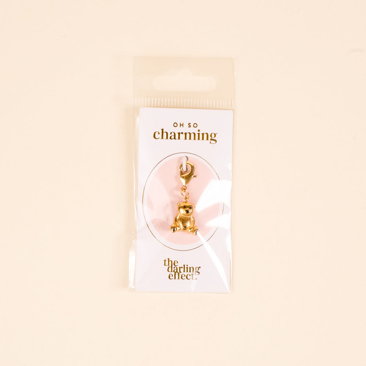 A gold teddy bear jewelry charm by The Darling Effect is attached to a card labeled OH SO charming and packaged with the card and charm bar in a clear plastic bag against a beige background.
