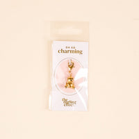A gold teddy bear jewelry charm by The Darling Effect is attached to a card labeled OH SO charming and packaged with the card and charm bar in a clear plastic bag against a beige background.