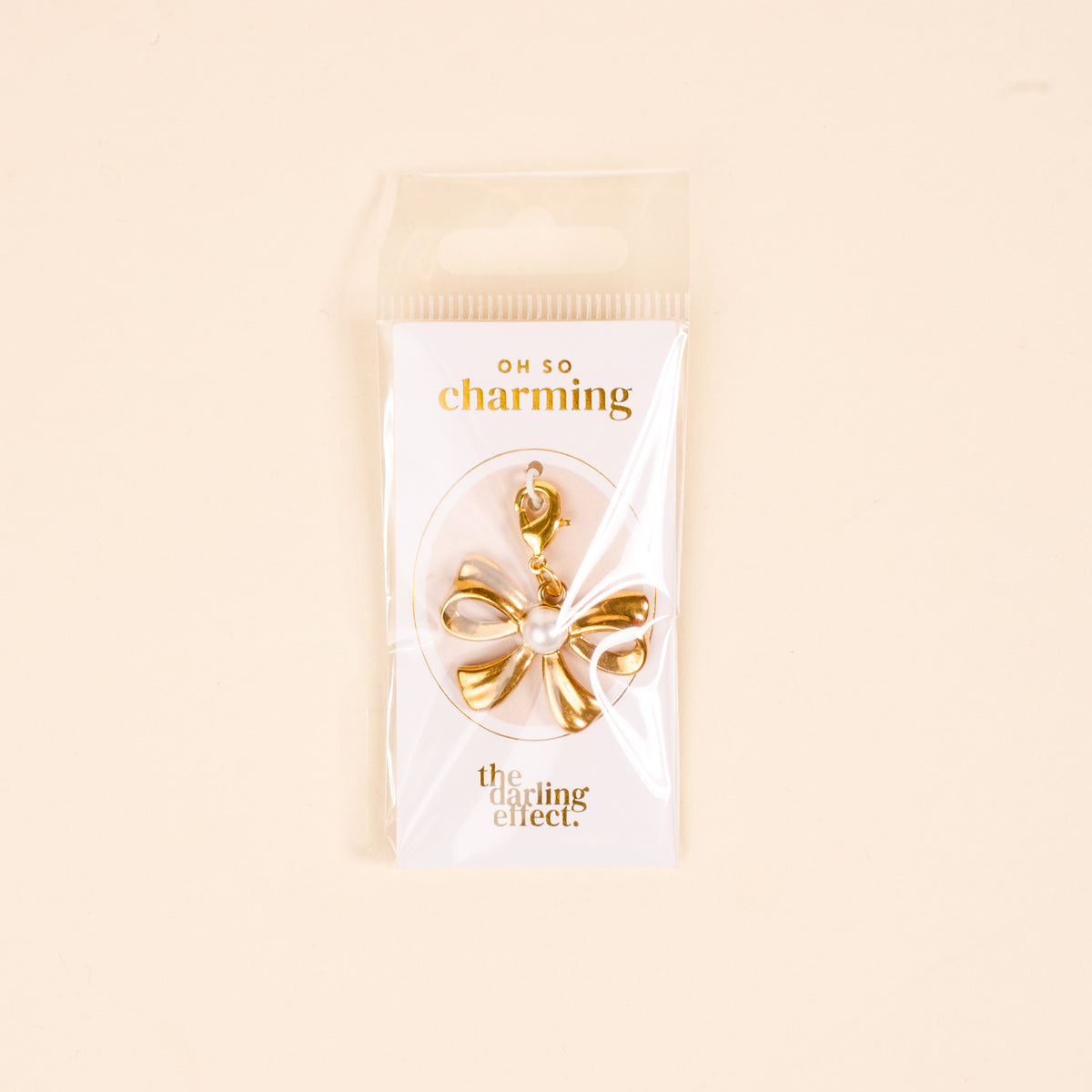 The Darling Effect's All Tied Up Jewelry Charm Gold features a gold and white bow shape with a clasp, displayed on an "OH SO charming" card against a light beige background.