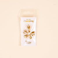 The Darling Effect's All Tied Up Jewelry Charm Gold features a gold and white bow shape with a clasp, displayed on an "OH SO charming" card against a light beige background.