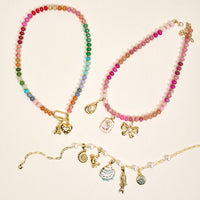 The Darling Effect’s Tulip Time Jewelry Charm Gold necklaces showcase vibrant beads and gold charms—heart, flower, bow, cupcake—each delicately detailed and artfully arranged on a cream background.