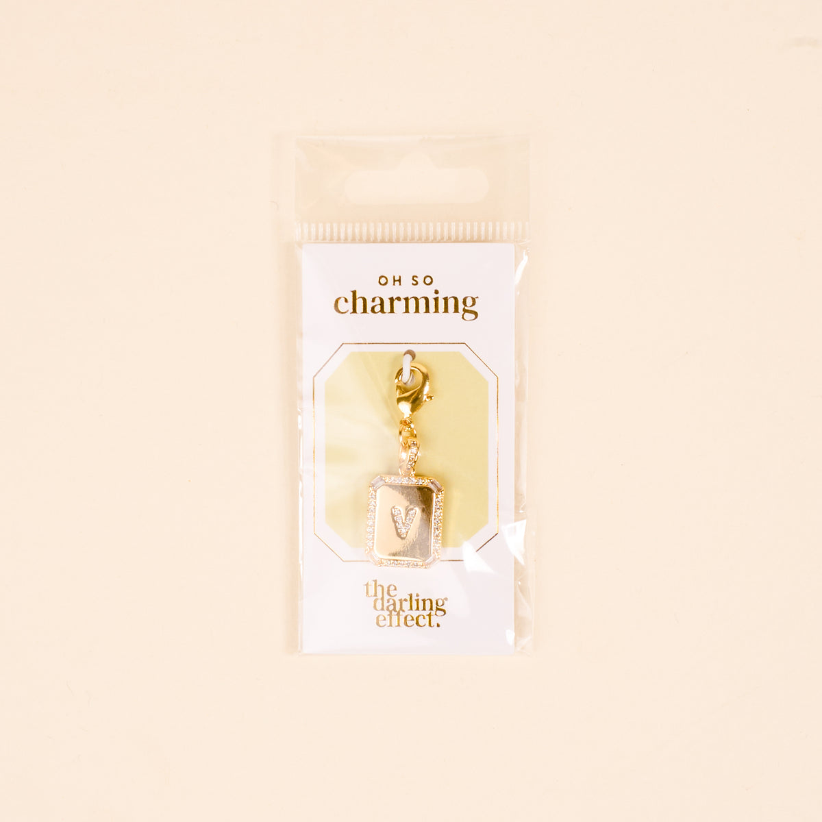 The-darling-effect's V Jewelry Charm Initial Letter Gold is a gold-plated rectangular pendant with the letter V, packaged in clear plastic—perfect for fans of personalized and Initial Letter Jewelry Charms.