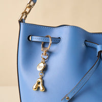 Close-up of a blue leather handbag with gold hardware and a drawstring, featuring the-darling-effect’s A Bubble Letter Keychain Bag Charm—a shiny gold personalized accessory with a small round tag.