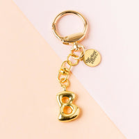 The B Bubble Letter Keychain Bag Charm by the-darling-effect features a gold bubble letter B and a round tag engraved with The Uniting Effect, set against a pastel pink and peach background.