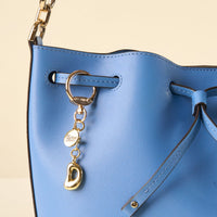 Close-up of a blue leather handbag with gold hardware and a drawstring closure, featuring the-darling-effect’s D Bubble Letter Keychain Bag Charm and a round engraved "by shine effect" accessory for personalized style.