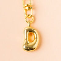 The-darling-effect D Bubble Letter Keychain Bag Charm features a shiny gold bubble letter "D" with a hollow bean-shaped charm, shown on a pale peach background.
