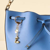 Close-up of a blue leather bucket bag with a gold chain strap and an E Bubble Letter Keychain Bag Charm by the-darling-effect, featuring a bubble "E" and branded disc.