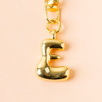 The E Bubble Letter Keychain Bag Charm by the-darling-effect features a shiny gold letter E on a chain against a light peach background, making it the perfect personalized accessory.