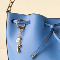 Close-up of a blue leather handbag with a drawstring closure, gold hardware, and the-darling-effect's F Bubble Letter Keychain Bag Charm hanging from the strap.