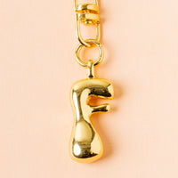 The-darling-effect's F Bubble Letter Keychain Bag Charm, a shiny gold charm in the shape of "F," hangs from a gold link on a light peach background—an ideal personalized accessory.