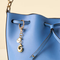A close-up of a blue leather handbag with black edges, adorned with the-darling-effect’s G Bubble Letter Keychain Bag Charm in gold and a small round tag on the strap; the bag features a drawstring closure.