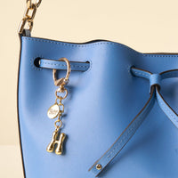 A close-up of a light blue leather handbag with a drawstring closure, featuring the-darling-effect's H Bubble Letter Keychain Bag Charm and a round Dior charm hanging from the strap.