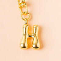 The H Bubble Letter Keychain Bag Charm by the-darling-effect features a bold, shiny gold letter H on a light peach background—a perfect personalized accessory for your bag.