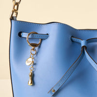 A close-up of a light blue leather bucket bag with gold hardware, a chain strap, and an I Bubble Letter Keychain Bag Charm by the-darling-effect for a unique touch.