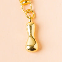 The I Bubble Letter Keychain Bag Charm by the-darling-effect features a shiny gold, bean-shaped pendant on a circular ring—an ideal personalized accessory for your bag, pictured here against a pale peach background.