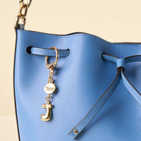 Close-up of a light blue leather bucket bag with a gold chain strap, styled with the-darling-effect J Bubble Letter Keychain Bag Charm, branded disc charm, and a drawstring closure.