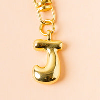 The-darling-effect’s J Bubble Letter Keychain Bag Charm features a shiny gold letter J pendant on a soft peach background—ideal for personalizing your keys or bag.