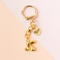 The-darling-effect K Bubble Letter Keychain Bag Charm features a gold, shiny bubble letter K and a round tag engraved with "life is calling collect," displayed on a pastel pink and peach background.