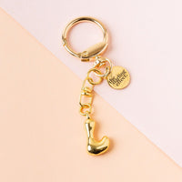 The-darling-effect's L Bubble Letter Keychain Bag Charm features a gold finish and a round tag engraved with "the gifting affair," perfect as a personalized bag charm, shown on a soft pink and peach background.