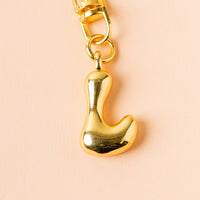 The-darling-effect's shiny, gold L Bubble Letter Keychain Bag Charm hangs from a clasp against a light peach background.