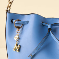 Close-up of the-darling-effect M Bubble Letter Keychain Bag Charm on a light blue leather handbag with a gold chain, personalized keychain, round "Be your own cheer" tag, and drawstring closure.