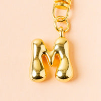 The-darling-effect's M Bubble Letter Keychain Bag Charm features a shiny gold, bubbly M with rounded edges and a gold clasp—ideal for personalizing your keys or bag.