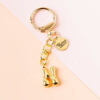 The-darling-effect’s N Bubble Letter Keychain Bag Charm features a shiny sculptural gold letter “N” and a round tag engraved with “the giving effect,” shown on a two-tone light peach and beige background.