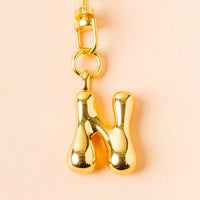 The-darling-effect N Bubble Letter Keychain Bag Charm features a chunky, shiny gold letter N pendant on a light peach background—ideal for personalizing keys or as a cute bag charm.