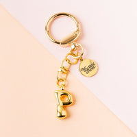 The P Bubble Letter Keychain Bag Charm by the-darling-effect features a gold letter P and a round tag engraved with the brand name, shown on a peach and light pink background.