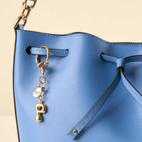 Close-up of a bright blue leather bucket bag featuring the-darling-effect’s P Bubble Letter Keychain Bag Charm in gold, hanging from a drawstring loop with a gold chain strap.