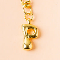 The-darling-effect P Bubble Letter Keychain Bag Charm features a shiny gold letter "P" pendant on a gold keychain, displayed on a peach background.