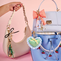 A hand holds a cream handbag with the-darling-effect’s P Bubble Letter Keychain Bag Charm, next to pastel purple and blue bags adorned with bows and beads, all styled against a pink background.