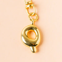 The Q Bubble Letter Keychain Bag Charm by the-darling-effect features a shiny gold finish, a circular hollow center, and a small bottom protrusion—perfect for personalizing your bag. Shown against a pale peach background.