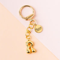 The R Bubble Letter Keychain Bag Charm by the-darling-effect features a shiny, rounded gold R charm and a small healing effect tag, displayed on a peach and cream background—perfect as a stylish accessory or thoughtful gift.