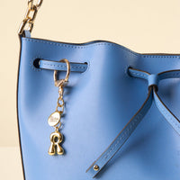 Close-up of a bright blue bucket bag with a gold chain strap and a gold keyring featuring the-darling-effect R Bubble Letter Keychain Bag Charm, with "darling effect" text and a golden R charm.