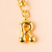 The-darling-effect's R Bubble Letter Keychain Bag Charm features a shiny gold, rounded R pendant—ideal as a personalized keychain or playful bag accessory. Displayed against a pale peach background.