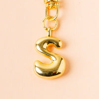 The S Bubble Letter Keychain Bag Charm by the-darling-effect features a shiny gold "S" pendant on a gold chain, displayed against a light peach background—ideal as a personalized accessory for your bag or keys.