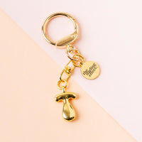 The T Bubble Letter Keychain Bag Charm by the-darling-effect is shown on a two-toned pastel background, featuring a gold mushroom-shaped charm and a round tag engraved with the brand name.