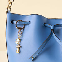 Close-up of a light blue leather handbag with drawstring closure and gold hardware, featuring the-darling-effect's T Bubble Letter Keychain Bag Charm—a playful accessory with a small round tag attached to the strap.