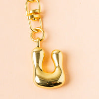 The U Bubble Letter Keychain Bag Charm by the-darling-effect features a shiny gold letter U bubble charm on a gold link, displayed on a light peach background—ideal for personalizing your bag.