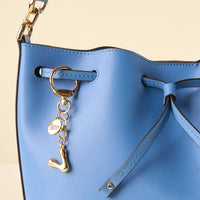 Close-up of a light blue leather bucket bag with a gold chain strap. Hanging from a grommet is the-darling-effect's V Bubble Letter Keychain Bag Charm, featuring a round gold charm with an abstract J.