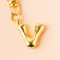 Close-up of the-darling-effect's V Bubble Letter Keychain Bag Charm, a shiny gold pendant with a clasp, perfect for adding personalized style to your bag, displayed against a soft pink background.