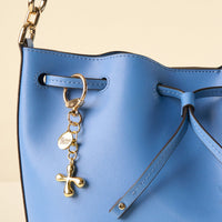A close-up of a blue leather drawstring bucket bag with a gold chain strap, featuring the-darling-effect’s X Bubble Letter Keychain Bag Charm and a round charm reading "the darling effect" alongside a chic gold cross.