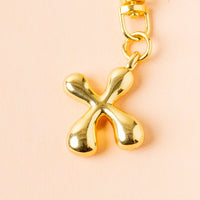 The X Bubble Letter Keychain Bag Charm by the-darling-effect, a shiny gold abstract accessory with rounded ends, hangs from a chain against a light peach background.